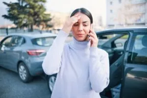 Woman wearing a turtleneck sweater with her cell phone in her left hand and her right hand on her forehead who is standing in front of two vehicles and trying to figure out who is liable for a Postmates car accident