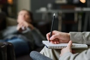 A therapist holds a notepad while a patient lies on the couch in the background and wonders about the signs of therapist sexual abuse. 