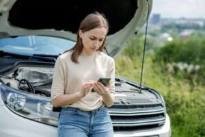 A woman on the phone next to a broken down car wondering if you can get a ticket days after an accident.