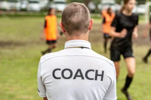 The back of a man wearing a white shirt with the word “Coach” written in black letters as he wonders if he can coach a youth sports team with a DUI.