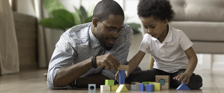 A father helping his child stack wooden toy blocks on the floor.