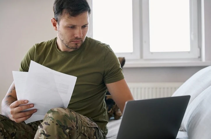 military member holding papers while looking at a laptop