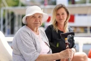 An elderly woman smiles as she sits on her patio, holding a cane. Her legal guardian, who gets paid in Virginia, sits with her.