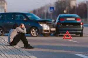 A man sits next to a car accident with his head in his hands.