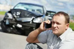 Man on his cell phone kneeling in front of two cars involved in a crash and wondering how long after a car accident they have to file a police report in NYC