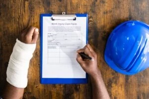 A person with a broken arm in a white cast is filling out a workers’ compensation form for pain and suffering in California next to a blue hard hat.