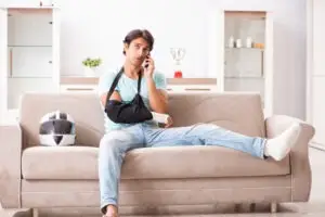 A motorcycle accident victim with his arm in a sling and leg in a cast sitting on a couch beside his helmet and calling an experienced motorcycle accident lawyer in Ventura.