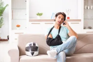 A motorcycle accident victim with his arm in a sling and foot in a cast sitting on a couch beside his helmet and calling an experienced motorcycle accident lawyer in Santa Ana.