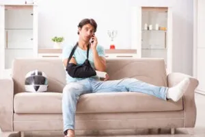 A motorcycle accident victim with his arm in a sling and foot in a cast sitting on a couch beside his helmet and calling an experienced motorcycle accident lawyer in Santa Barbara.