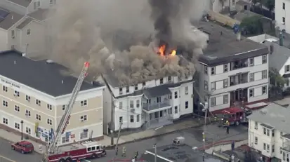Aerial view of a multi-story residential building engulfed in flames with heavy smoke, as firefighters on the ground and on an extended ladder battle the blaze
