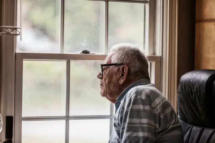 An elderly man with glasses, wearing a striped shirt, gazes out of a window with a contemplative expression.