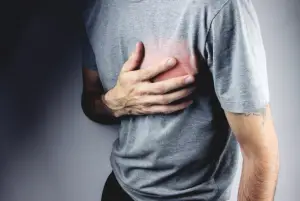 Person clutching their chest in discomfort, with a visible red area suggesting pain or injury, on a grey background