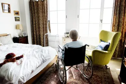 An elderly person in a wheelchair facing a bright window in a cozy bedroom