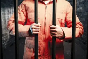A prisoner in an orange jumpsuit holds prison bars.
