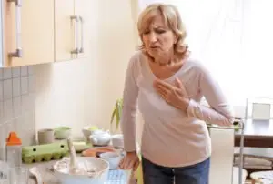 A woman in her kitchen grips at her chest.