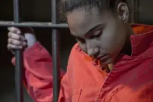 A woman in a prison jumpsuit holding cell bars.