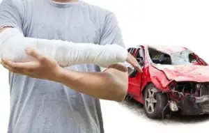 A man with a bandaged arm near a wrecked car.