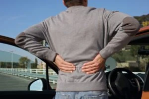 A man grasping his back in pain outside of his red car.