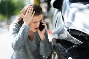 A woman on her phone, looking that the damage caused to her vehicle by a car accident.