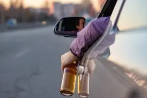 A man’s arm hanging out the driver’s side window of a car holding a bottle of alcohol.
