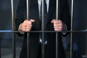 A man in a black suit is in jail with his hands on the bars.