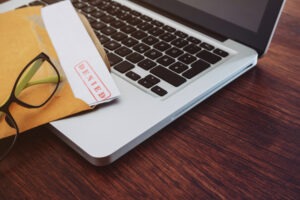 An overhead view of a white piece of paper with a red “claim denied” stamp peeking out of a brown envelope and a pair of glasses on top of a laptop keyboard.