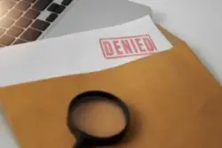 A white paper with a red “claim denied” stamp peeking out of a brown envelope on a laptop computer and a magnifying glass on top.